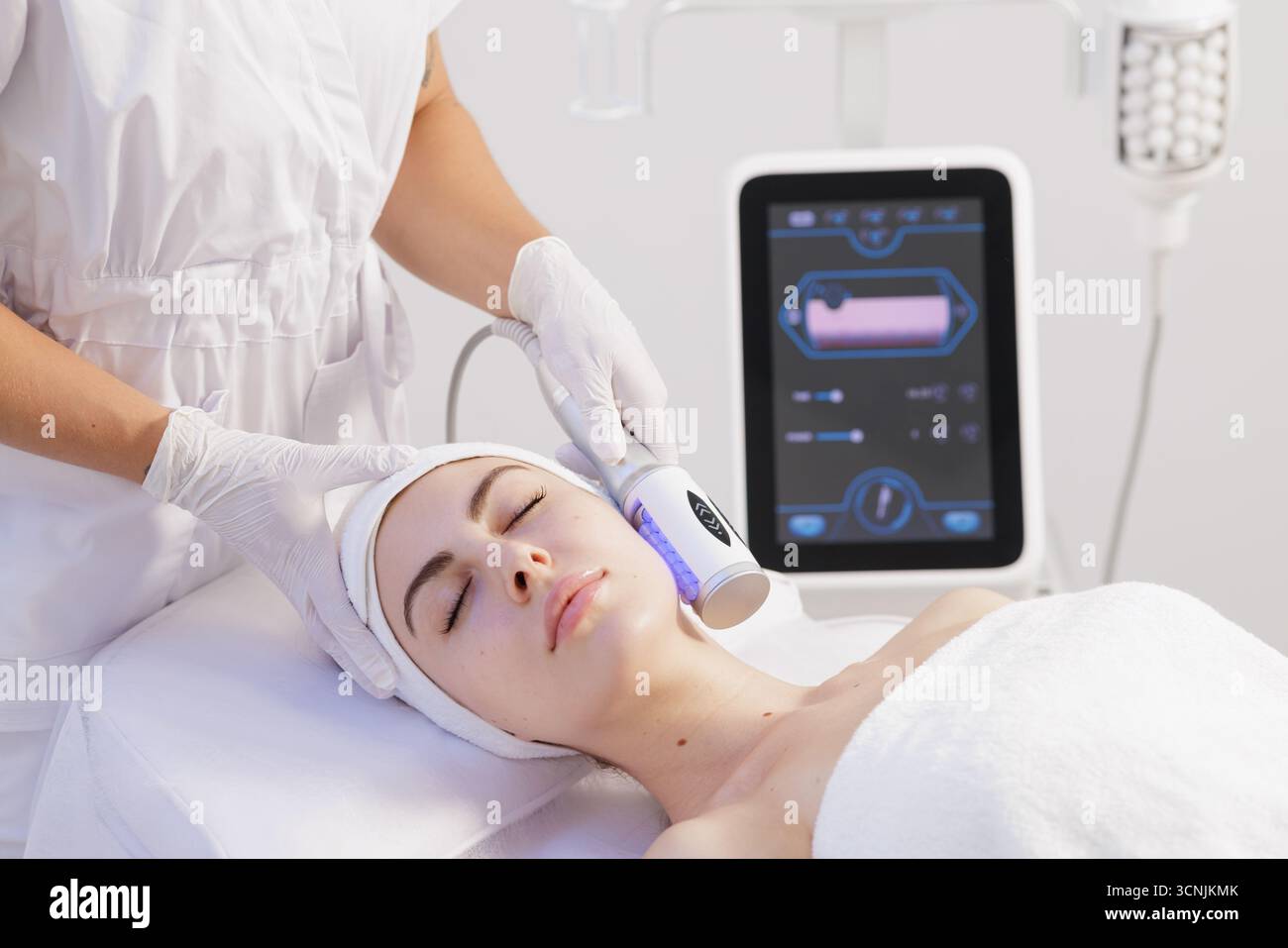 Le praticien ganté utilise un rouleau de microvibration sur un visage de client détendu dans une clinique lumineuse pour soulever, contouring et tonifier la peau. Banque D'Images