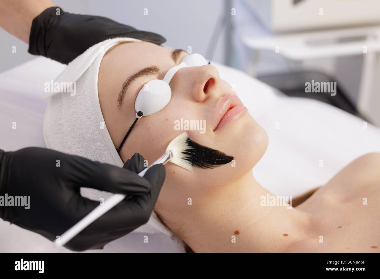 Esthéticienne en gants applique une peau noire avec une brosse sur le visage d'une femme avec des boucliers oculaires dans une clinique lumineuse pour l'exfoliation et le renouvellement de la peau. Banque D'Images