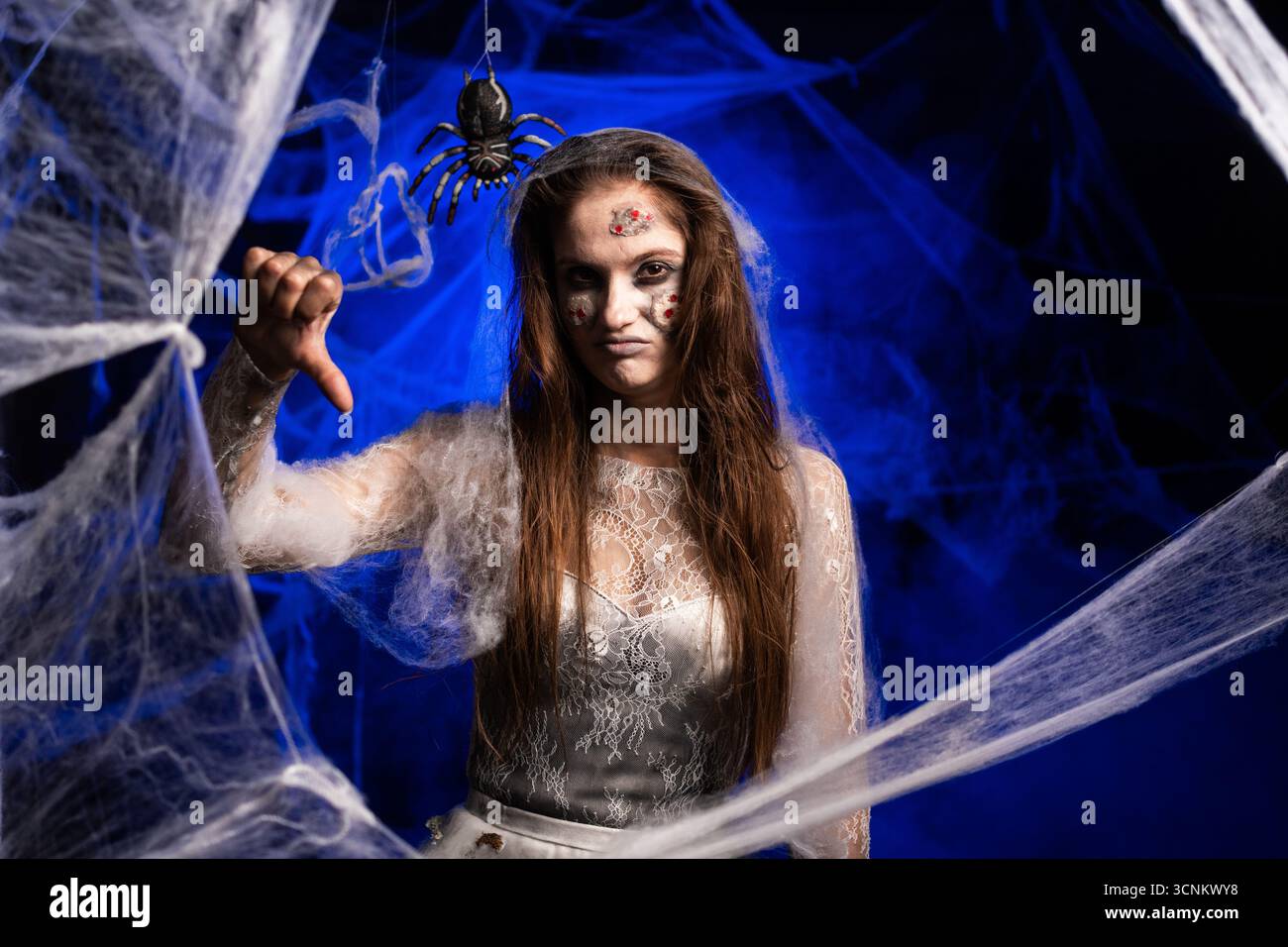 Malheureuse femme effrayante avec horrible maquillage de zombie effrayant Halloween en robe blanche montrant le geste détester pouces-bas debout sur fond bleu surrou Banque D'Images