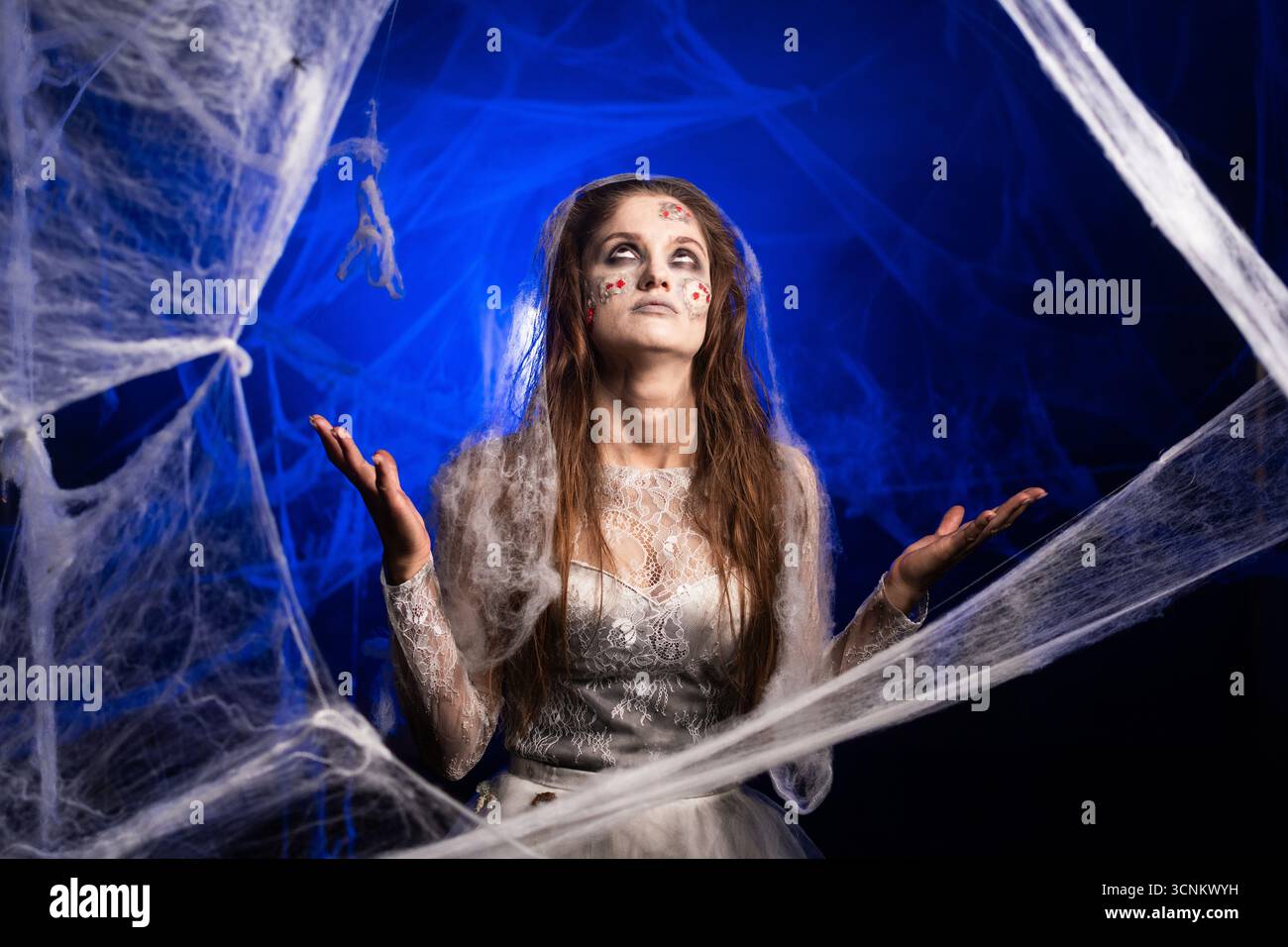 Stressé fatigué femme effrayante avec horrible maquillage zombie effrayant Halloween en robe blanche debout sur fond bleu entouré de toile d'araignée. Devil Obsesse Banque D'Images
