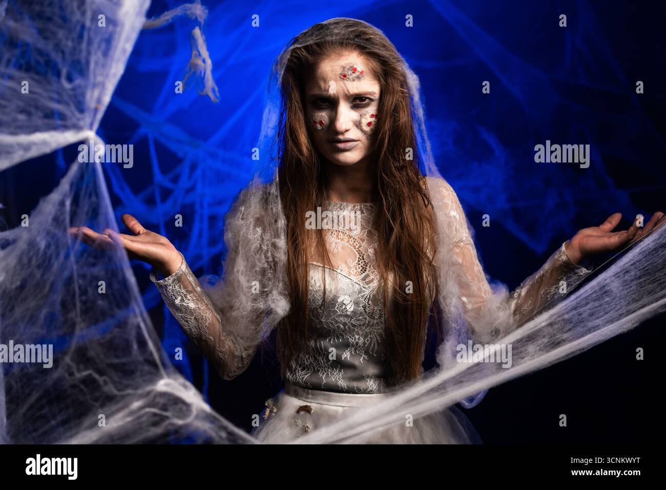 Malheureuse femme effrayante avec horrible maquillage zombie effrayant Halloween en robe blanche dit pourquoi voulez-vous, debout sur fond bleu entouré de toile d'araignée Banque D'Images