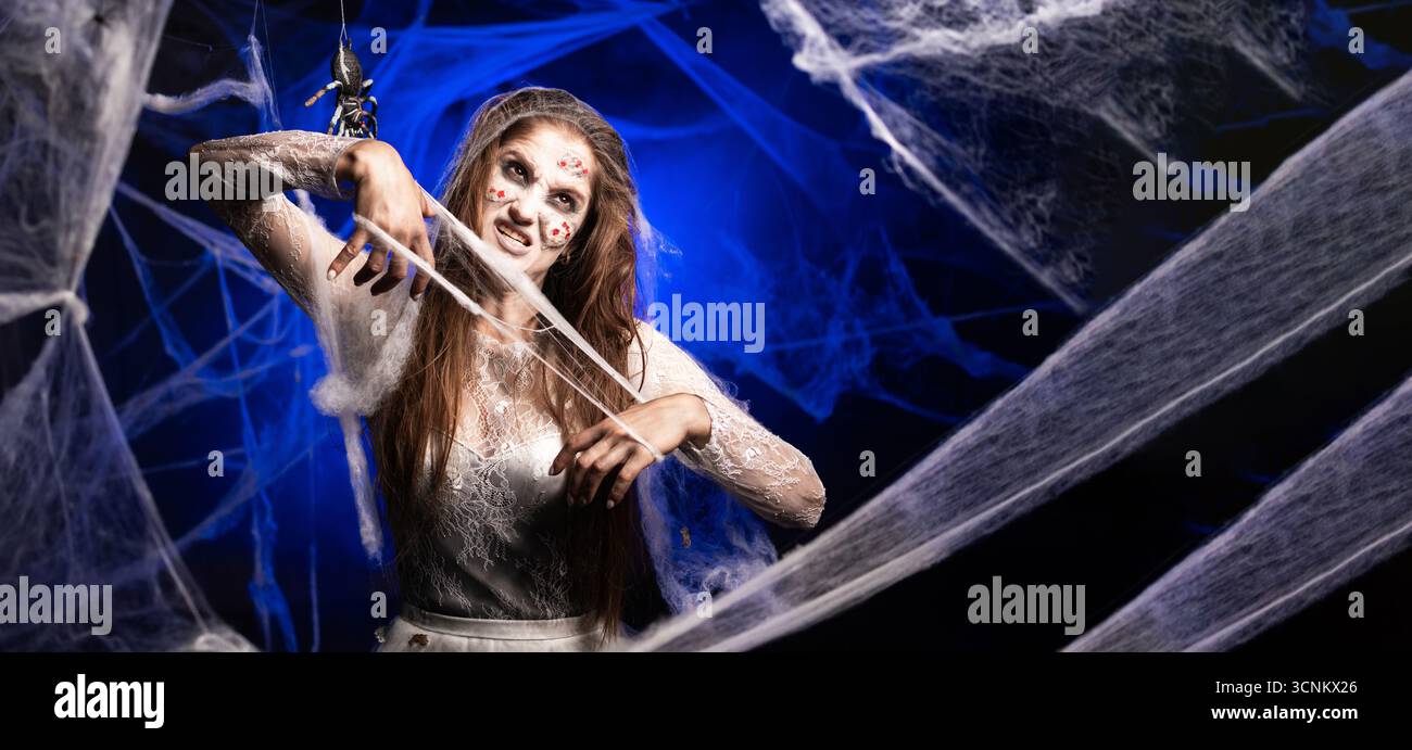 Femme effrayante avec horrible maquillage zombie effrayant Halloween en robe blanche déchirant la toile avec ses mains debout sur fond bleu entouré sp Banque D'Images