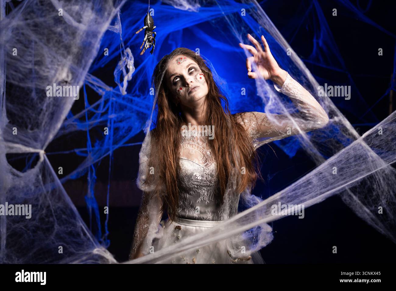 Sinistre femme effrayante avec horrible maquillage zombie effrayant Halloween en robe blanche montrant le geste OK debout sur fond bleu entouré de toile d'araignée. Banque D'Images