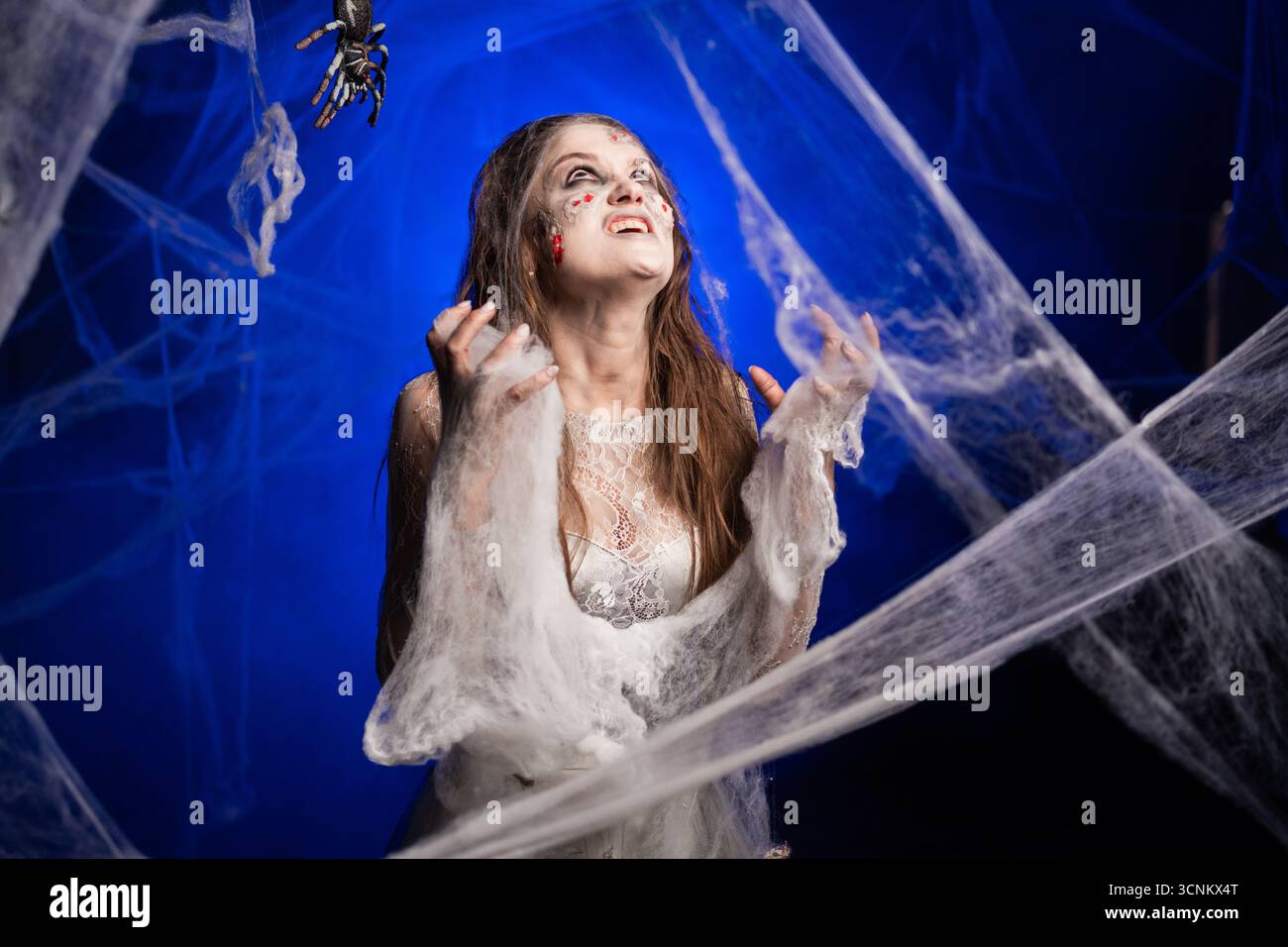 Femme effrayante avec horrible maquillage zombie effrayant Halloween en robe blanche criant avec des émotions agressives debout sur fond bleu entouré spid Banque D'Images