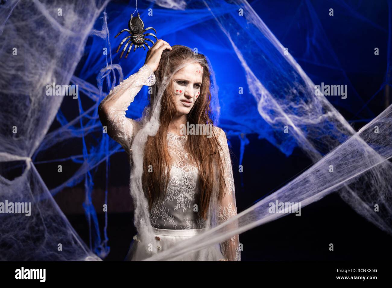 Triste femme sinistre confuse et perplexe avec le maquillage zombie effrayant Halloween tenir la tête à gratter. Fille morte avec le visage de cicatrices sur fond bleu de toile d'araignée. cop Banque D'Images