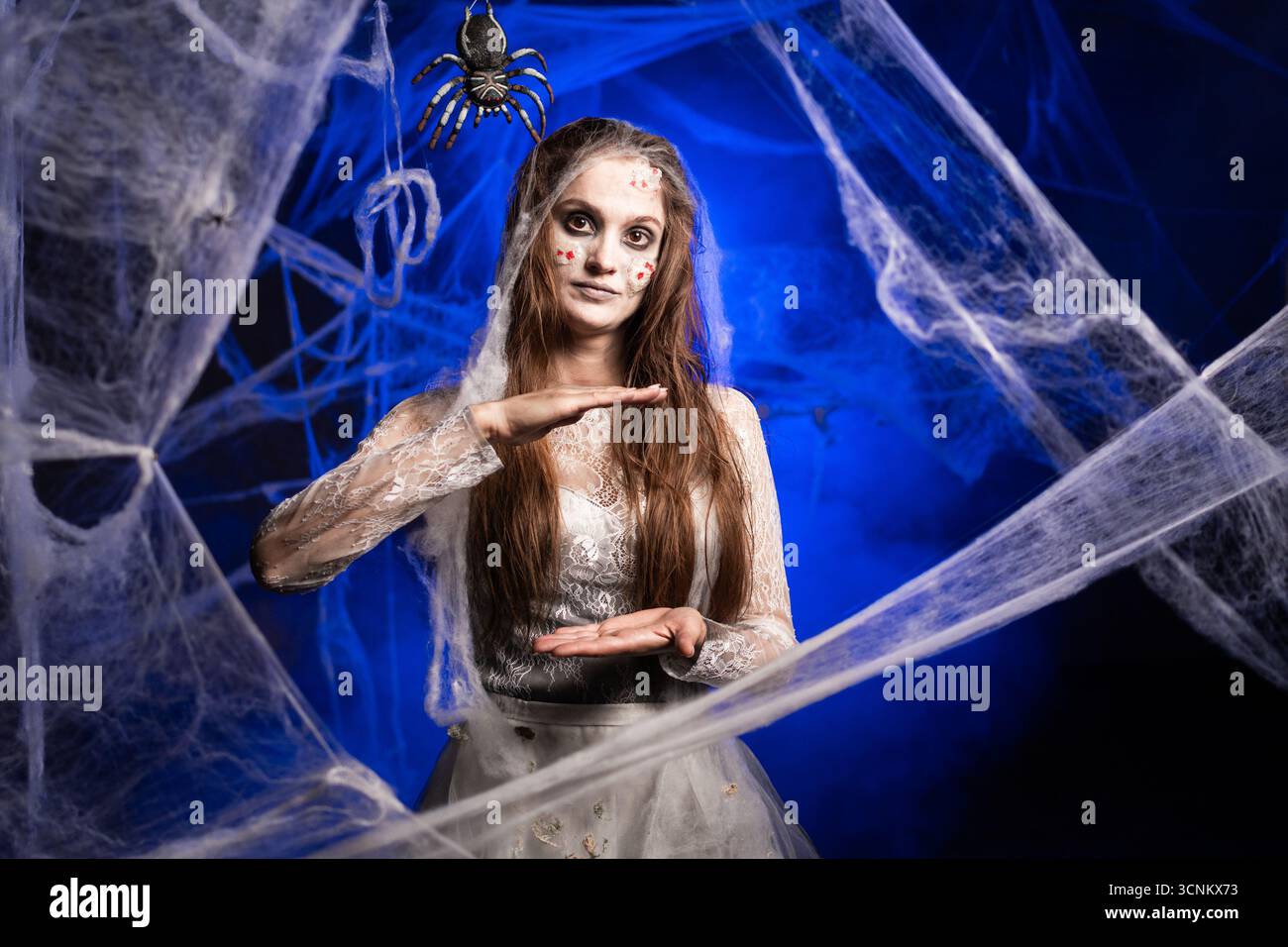 Femme sinistre dans une robe sale blanche avec un horrible maquillage zombie Halloween montrant un geste de grande taille avec ses doigts debout sur fond bleu surrou Banque D'Images