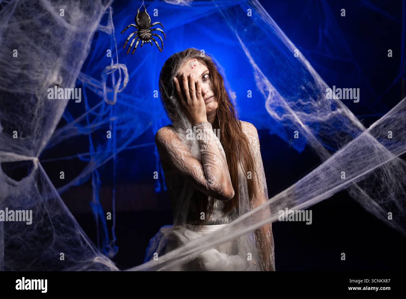 Triste femme sinistre avec le maquillage zombie effrayant d'Halloween mis la main sur le visage échouer un geste omg erroné debout sur fond bleu entouré de toile d'araignée. Banque D'Images