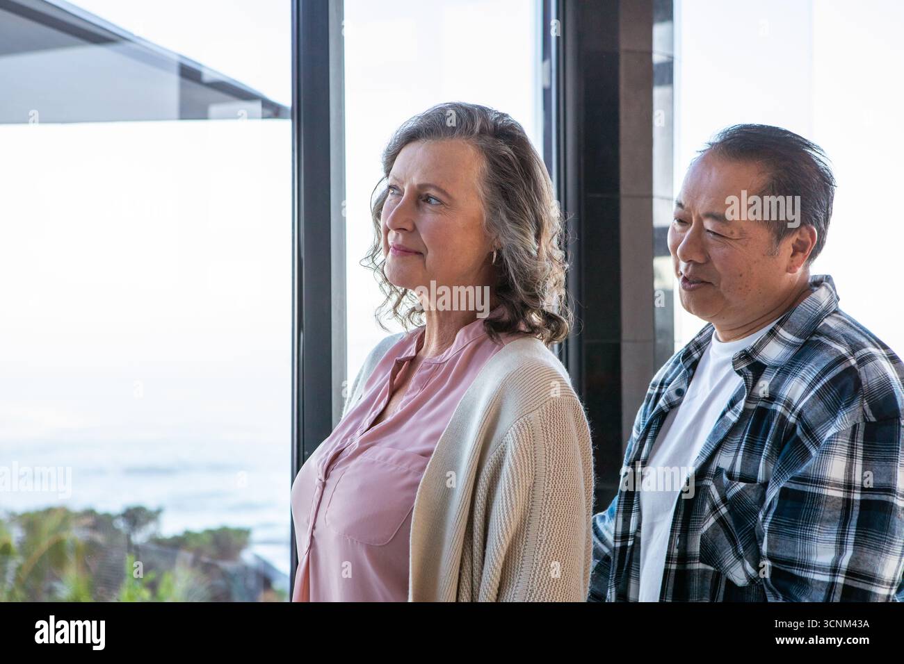 Couple senior diversifié debout regardant l'océan à travers la fenêtre à l'intérieur de la maison avec des plantes côtières Banque D'Images