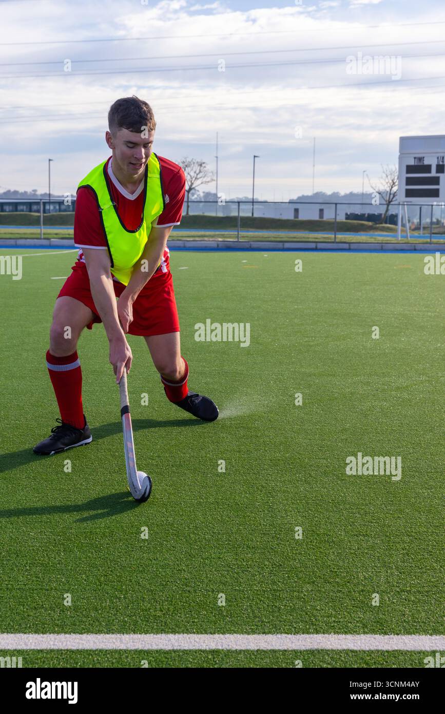 Joueur masculin de hockey sur gazon en rouge uniforme pliant et contrôlant la balle avec bâton sur le gazon Banque D'Images