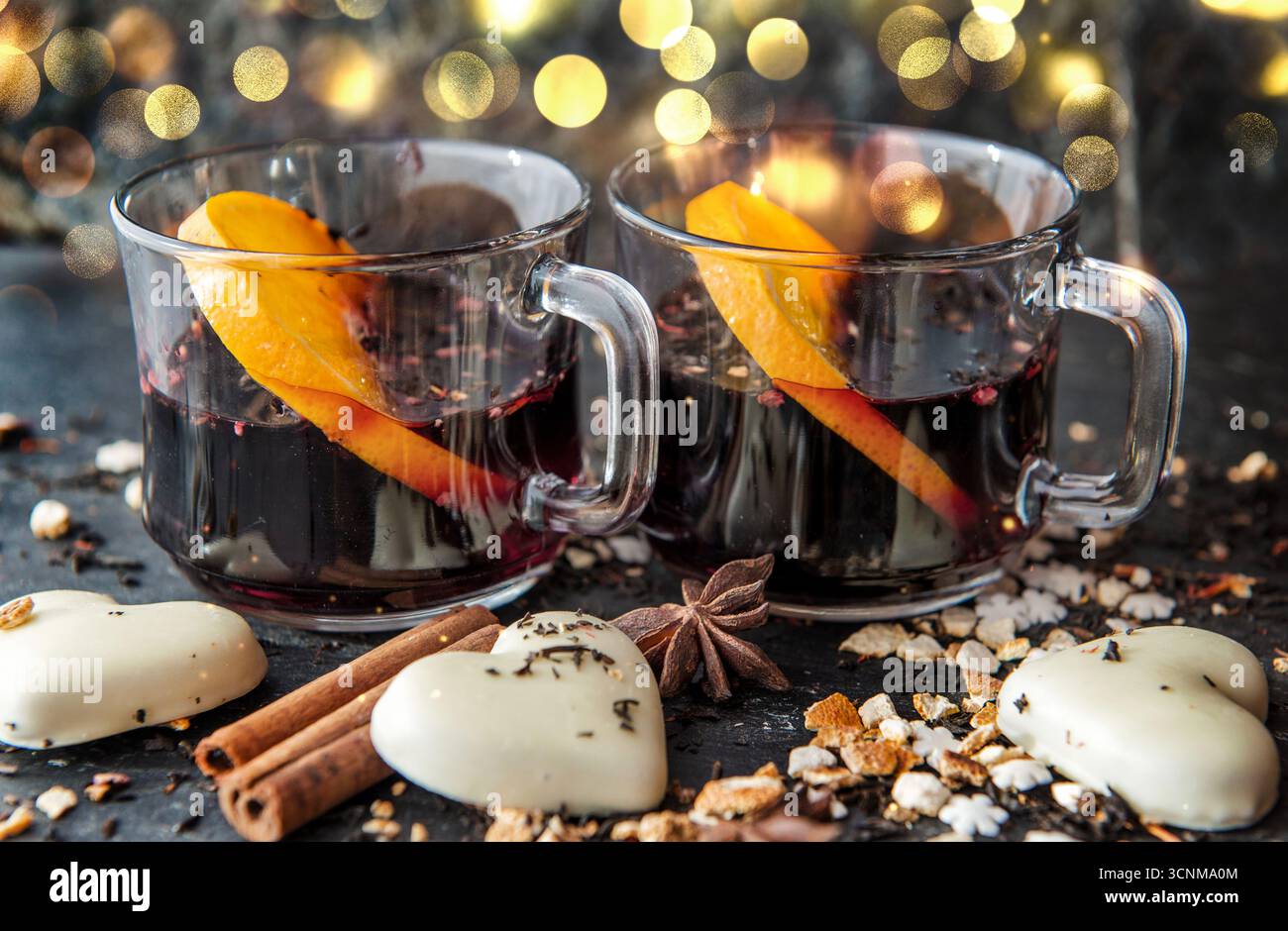tranche d'orange dans du vin chaud dans deux verres avec des biscuits en forme de coeur et des épices disposés sur fond de lumières floues Banque D'Images