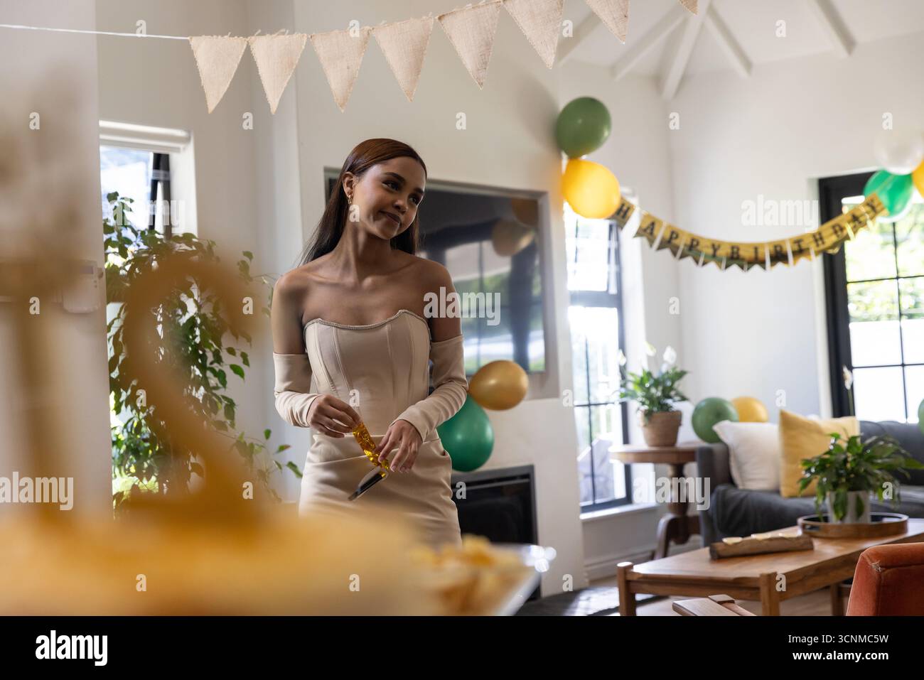 Femme asiatique portant une robe tenant corne de fête en or à la table de gâteau, ballons dans le salon Banque D'Images