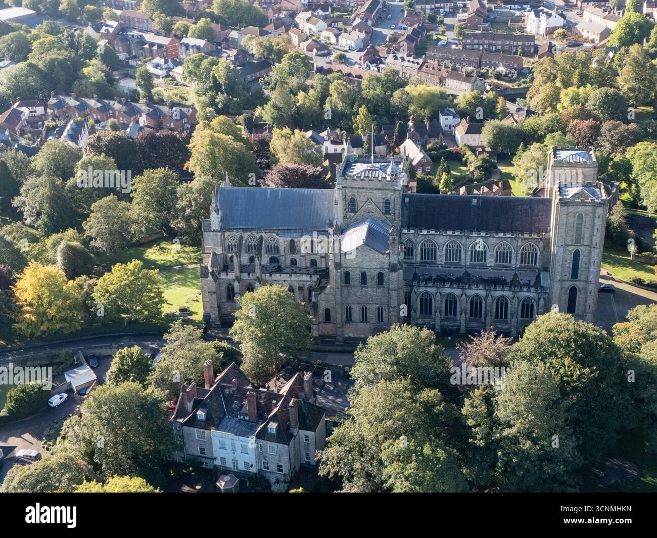 Photographie aérienne de la cathédrale de Ripon à Ripon, Yorkshire du Nord, Royaume-Uni, capturée par temps clair de fin d'été avec un ciel bleu vif et un soleil chaud mettant en valeur l'architecture gothique saisissante de la cathédrale. L'image montre l'élégant front ouest anglais ancien avec ses tours jumelles, ses pierres complexes et ses fenêtres cintrées, entouré par les rues historiques de la ville, les bâtiments voisins, les toits et les parcelles d'espaces verts, y compris les parcs et les espaces ouverts. Cette vue aérienne d'un drone capture la position centrale de la cathédrale dans la ville médiévale compacte, soulignant son rôle en tant que monument historique clé Banque D'Images