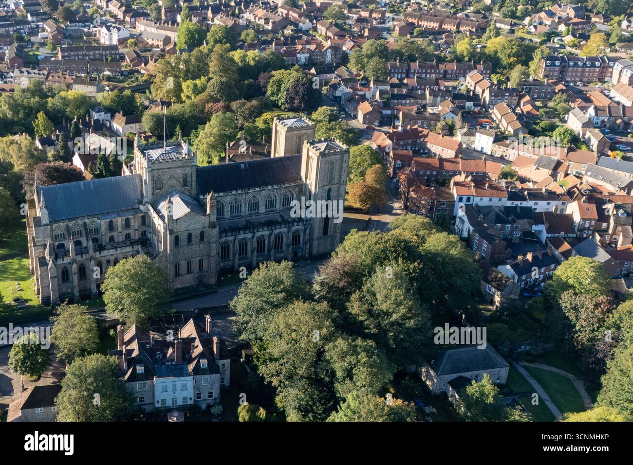 Photographie aérienne de la cathédrale de Ripon à Ripon, Yorkshire du Nord, Royaume-Uni, capturée par temps clair de fin d'été avec un ciel bleu vif et un soleil chaud mettant en valeur l'architecture gothique saisissante de la cathédrale. L'image montre l'élégant front ouest anglais ancien avec ses tours jumelles, ses pierres complexes et ses fenêtres cintrées, entouré par les rues historiques de la ville, les bâtiments voisins, les toits et les parcelles d'espaces verts, y compris les parcs et les espaces ouverts. Cette vue aérienne d'un drone capture la position centrale de la cathédrale dans la ville médiévale compacte, soulignant son rôle en tant que monument historique clé Banque D'Images