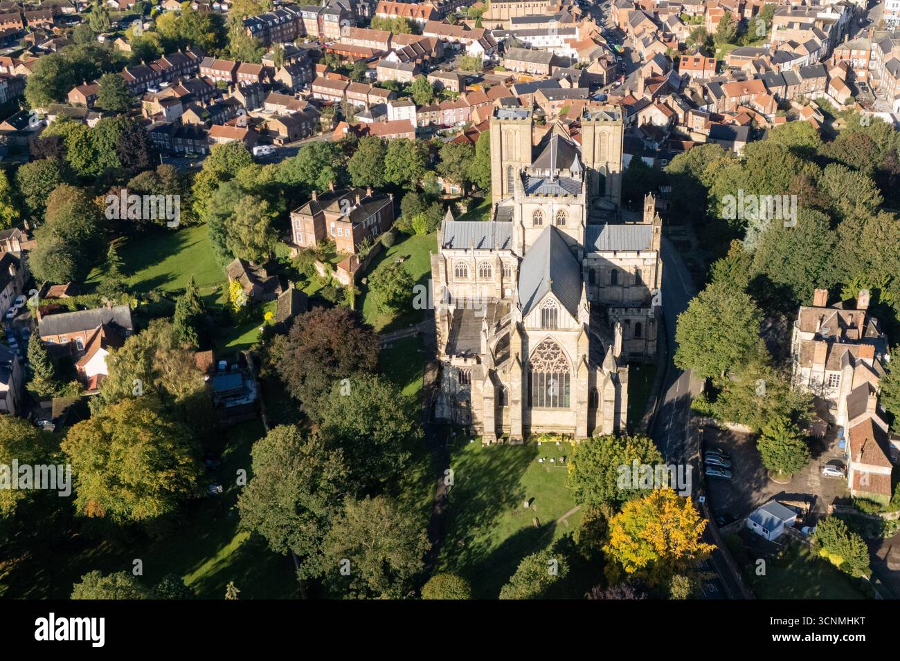 Photographie aérienne de la cathédrale de Ripon à Ripon, Yorkshire du Nord, Royaume-Uni, capturée par temps clair de fin d'été avec un ciel bleu vif et un soleil chaud mettant en valeur l'architecture gothique saisissante de la cathédrale. L'image montre l'élégant front ouest anglais ancien avec ses tours jumelles, ses pierres complexes et ses fenêtres cintrées, entouré par les rues historiques de la ville, les bâtiments voisins, les toits et les parcelles d'espaces verts, y compris les parcs et les espaces ouverts. Cette vue aérienne d'un drone capture la position centrale de la cathédrale dans la ville médiévale compacte, soulignant son rôle en tant que monument historique clé Banque D'Images