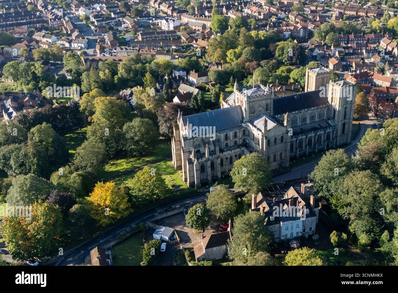 Photographie aérienne de la cathédrale de Ripon à Ripon, Yorkshire du Nord, Royaume-Uni, capturée par temps clair de fin d'été avec un ciel bleu vif et un soleil chaud mettant en valeur l'architecture gothique saisissante de la cathédrale. L'image montre l'élégant front ouest anglais ancien avec ses tours jumelles, ses pierres complexes et ses fenêtres cintrées, entouré par les rues historiques de la ville, les bâtiments voisins, les toits et les parcelles d'espaces verts, y compris les parcs et les espaces ouverts. Cette vue aérienne d'un drone capture la position centrale de la cathédrale dans la ville médiévale compacte, soulignant son rôle en tant que monument historique clé Banque D'Images