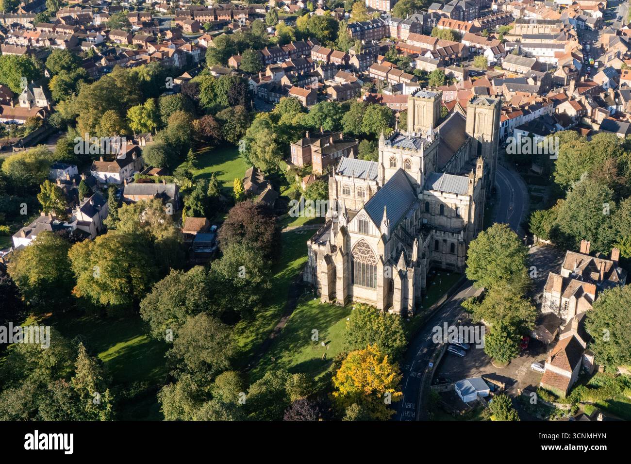Photographie aérienne de la cathédrale de Ripon à Ripon, Yorkshire du Nord, Royaume-Uni, capturée par temps clair de fin d'été avec un ciel bleu vif et un soleil chaud mettant en valeur l'architecture gothique saisissante de la cathédrale. L'image montre l'élégant front ouest anglais ancien avec ses tours jumelles, ses pierres complexes et ses fenêtres cintrées, entouré par les rues historiques de la ville, les bâtiments voisins, les toits et les parcelles d'espaces verts, y compris les parcs et les espaces ouverts. Cette vue aérienne d'un drone capture la position centrale de la cathédrale dans la ville médiévale compacte, soulignant son rôle en tant que monument historique clé Banque D'Images
