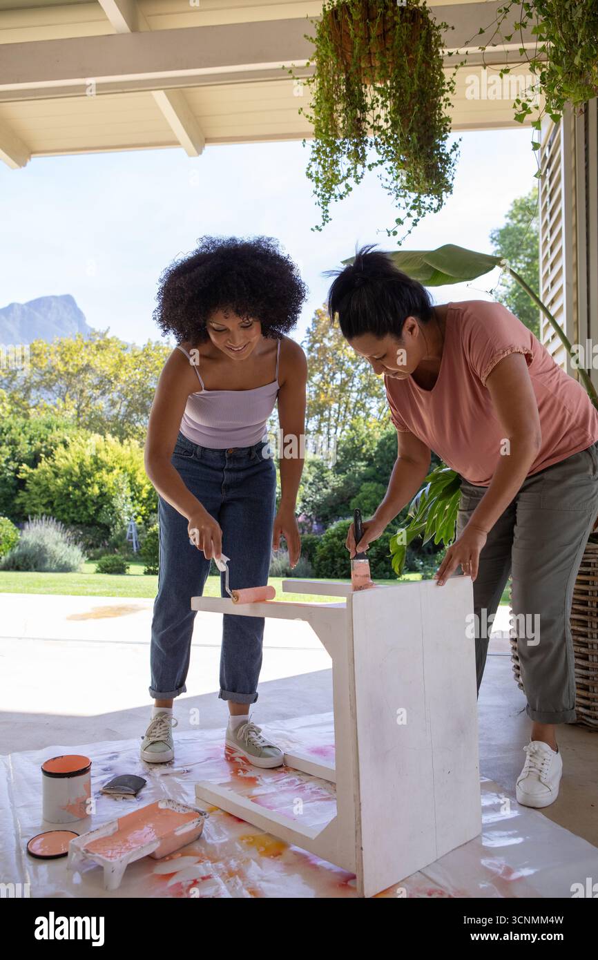 Mère diversifiée avec fille adulte peignant la table en bois avec de la peinture pêche sur le tissu de goutte sous le patio Banque D'Images