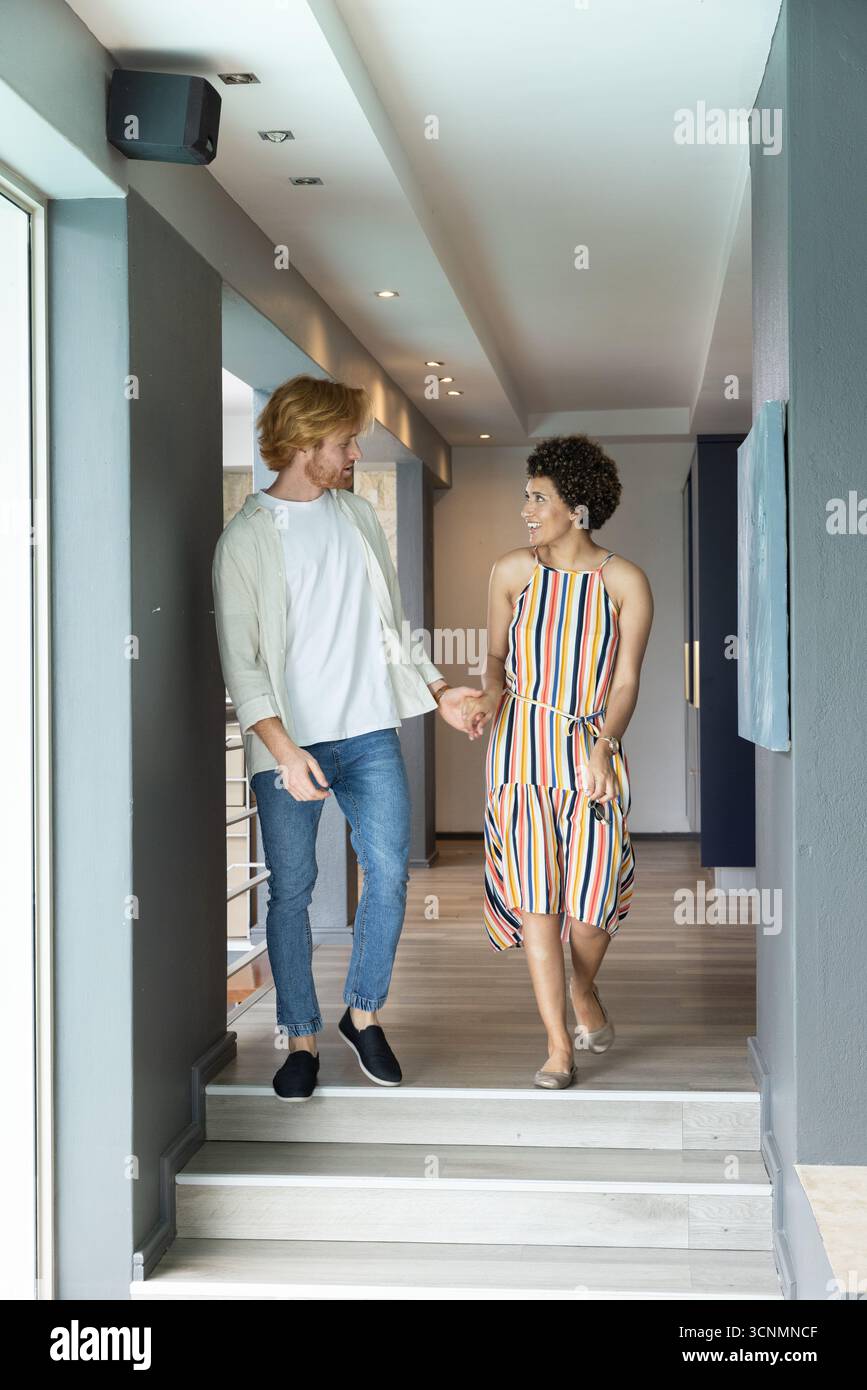 Couple diversifié marchant dans le vol des marches dans le couloir de la maison se tenant les mains Banque D'Images