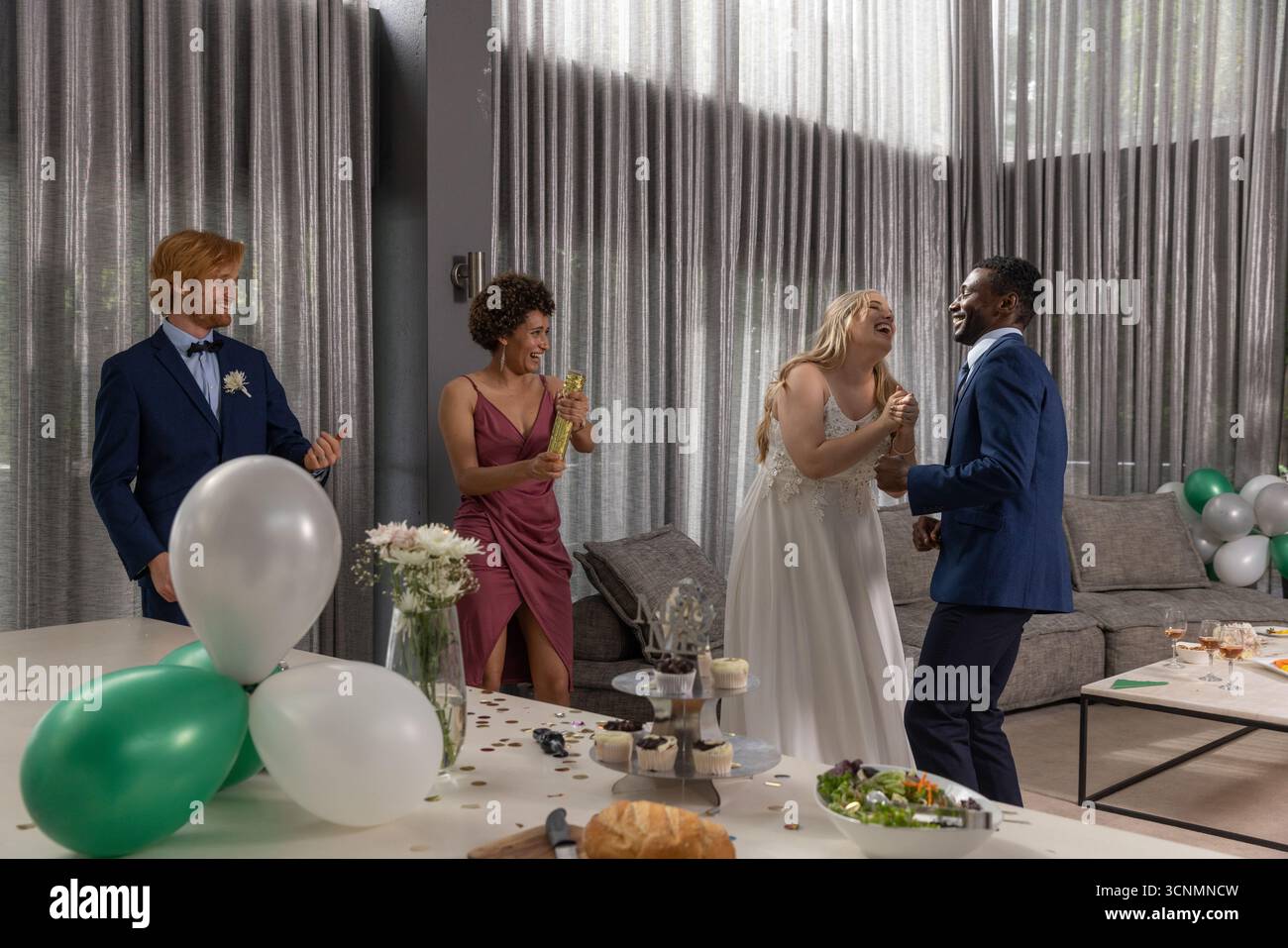 Couple diversifié dansant et riant dans la salle de réception décorée avec des ballons, stand de cupcake Banque D'Images