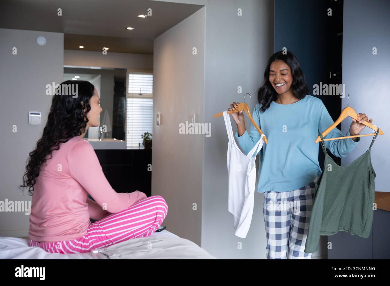 Amies féminines dans la chambre tenant des camisoles blanches et vertes sur des cintres comparant Banque D'Images Amies féminines dans la chambre tenant des camisoles blanches et vertes sur des cintres comparant Banque D'Images