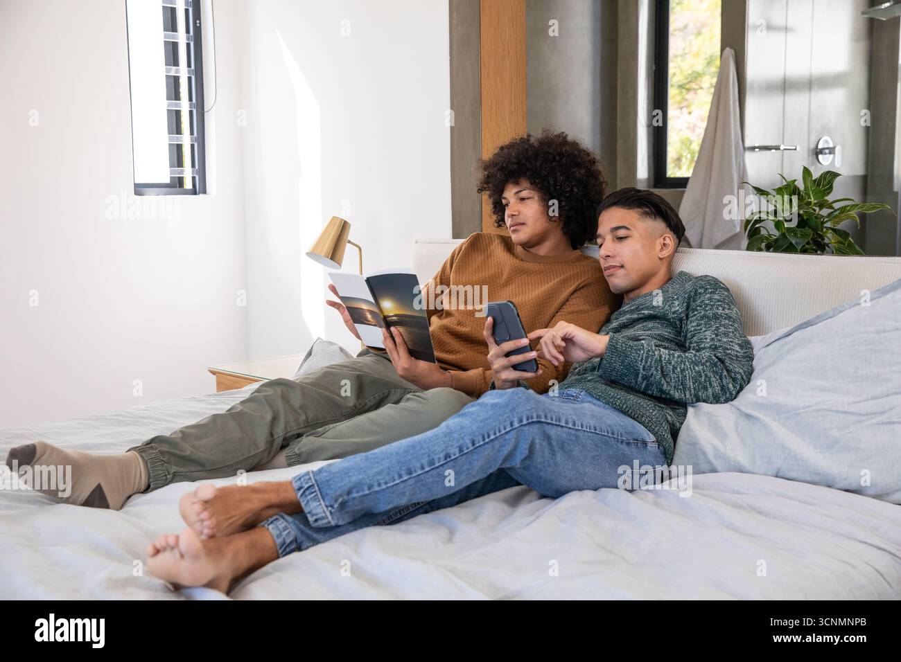 Amis afro-américains se relaxant sur le lit dans la chambre moderne lire magazine et à l'aide d'un smartphone Banque D'Images