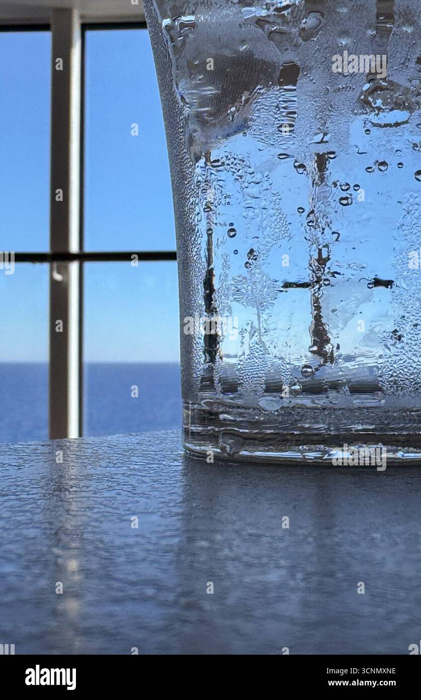 Un verre transparent avec condensation d'eau placé sur une surface réfléchissante, placé contre un horizon océanique tranquille et un ciel bleu visible à travers un windo encadré Banque D'Images