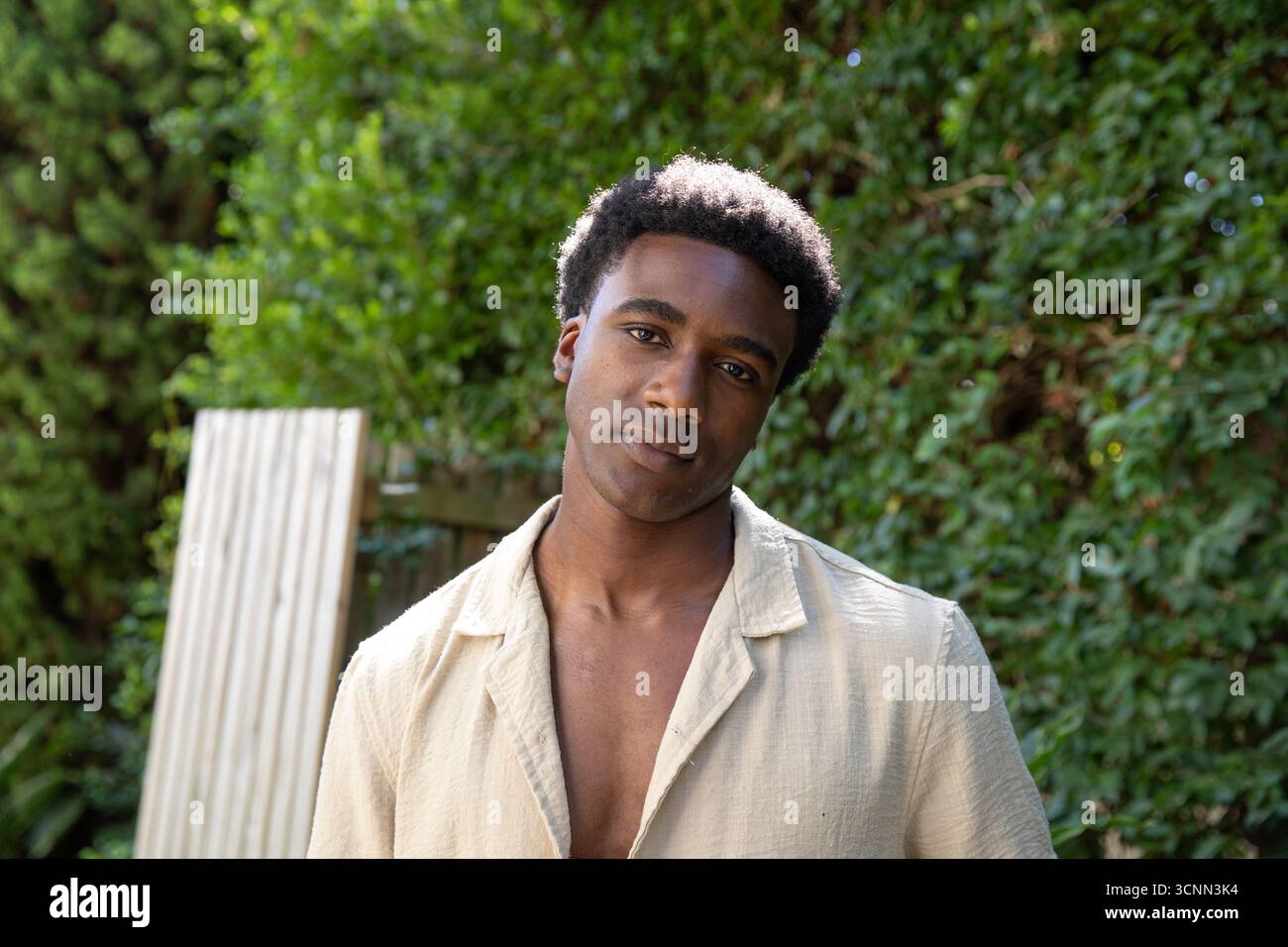 Homme afro-américain debout à l'extérieur dans la cour arrière par la planche de bois et la clôture à côté de la haie verte Banque D'Images