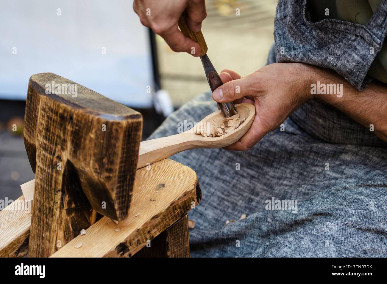 Gros plan d'un artisan sculptant une cuillère en bois à la main à l'aide d'outils traditionnels de travail du bois. Artisan en tablier travaillant avec du bois, copeaux visibles Banque D'Images
