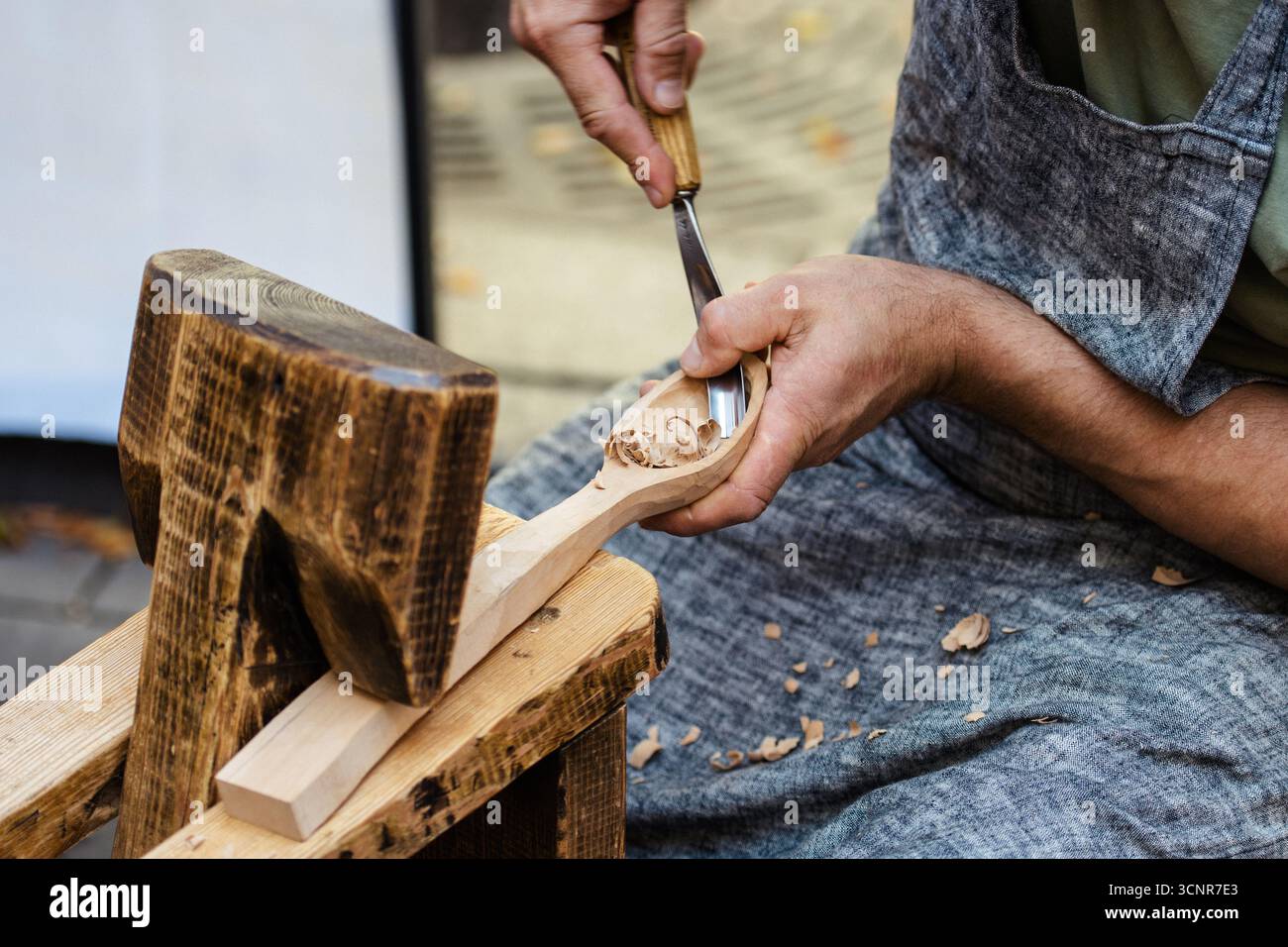 Gros plan d'un artisan sculptant une cuillère en bois à la main à l'aide d'outils traditionnels de travail du bois. Artisan en tablier travaillant avec du bois, copeaux visibles Banque D'Images