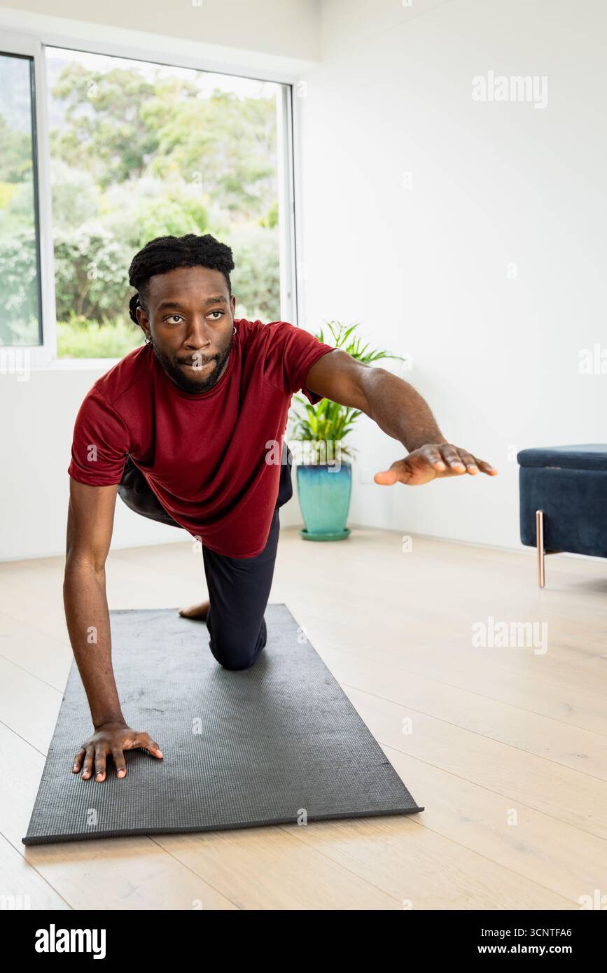 Homme afro-américain équilibrant sur tapis de yoga noir à la maison avec plante feuillue et fenêtre Banque D'Images
