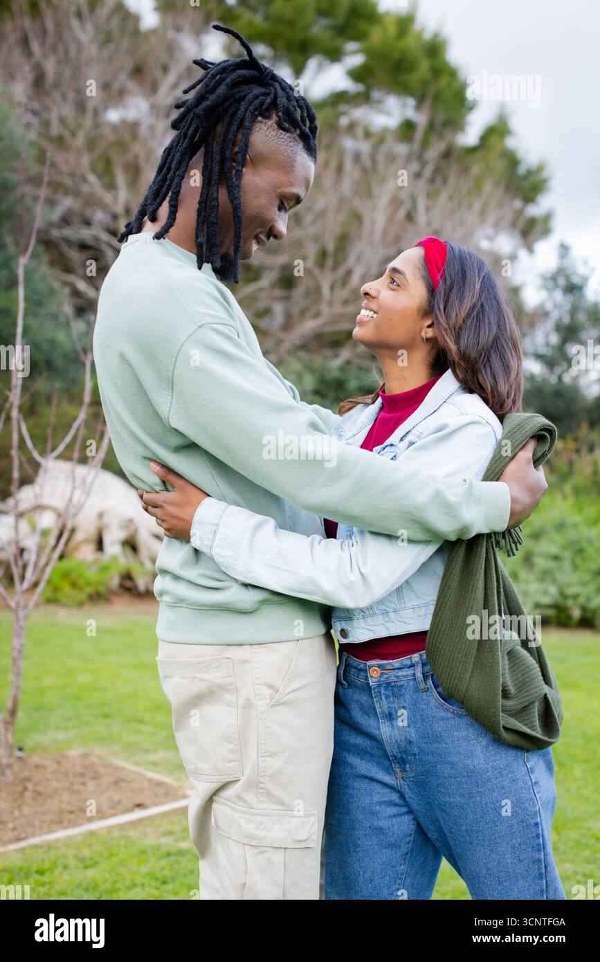 Couple diversifié embrassant sur la pelouse herbeuse dans le parc portant une veste en denim et tenant un pull vert Banque D'Images