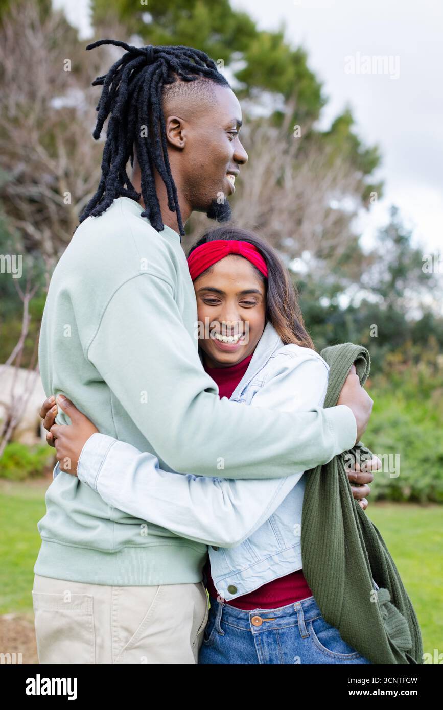 Couple diversifié embrassant dans un parc herbeux avec veste en denim et bandeau rouge tenant un pull en tricot vert Banque D'Images