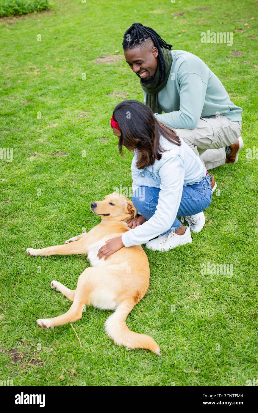 Couple indien et afro-américain agenouillé et caressant Golden retriever sur un champ herbeux dans le parc Banque D'Images
