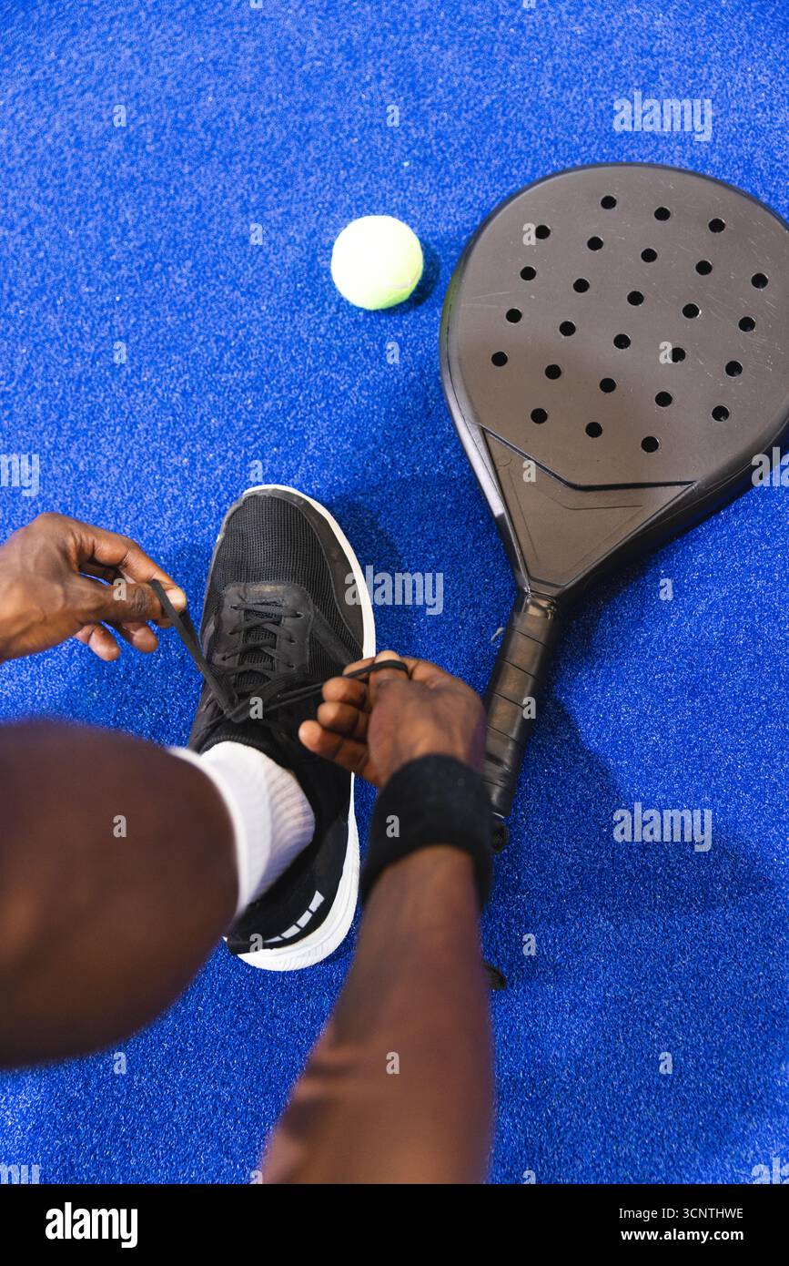Homme afro-américain attachant des lacets noirs sur un terrain de padel avec une raquette de padel et une balle jaune-vert. Athlète, sports, équipement, chaussures, fitness, acti Banque D'Images