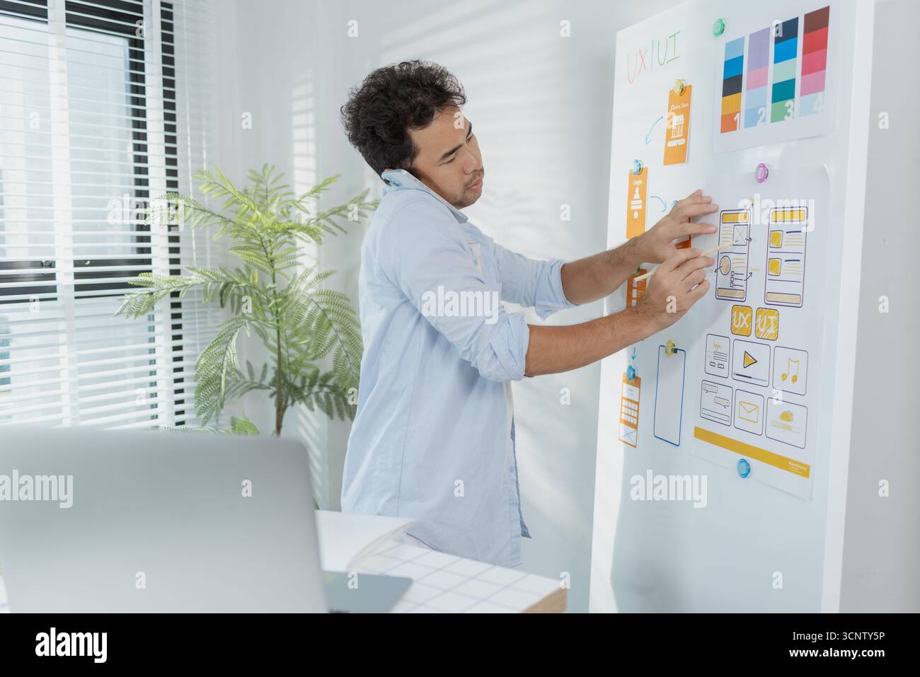 Un concepteur créatif travaille sur la planification UX/UI, analysant des croquis et des prototypes sur un tableau blanc tout en utilisant un ordinateur portable. Le processus montre la pensée de conception Banque D'Images