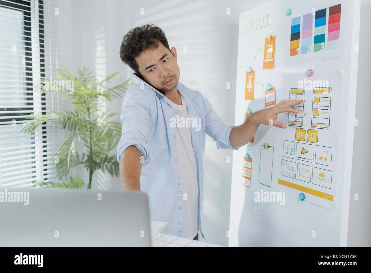 Un concepteur créatif travaille sur la planification UX/UI, analysant des croquis et des prototypes sur un tableau blanc tout en utilisant un ordinateur portable. Le processus montre la pensée de conception Banque D'Images