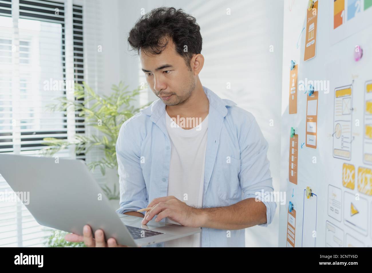Un concepteur créatif travaille sur la planification UX/UI, analysant des croquis et des prototypes sur un tableau blanc tout en utilisant un ordinateur portable. Le processus montre la pensée de conception Banque D'Images