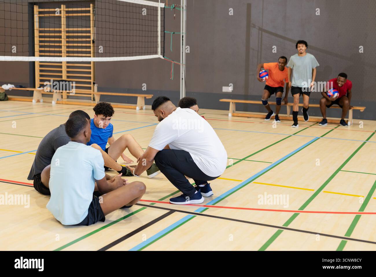 Entraîneur afro-américain agenouillé sur un terrain de volley-ball de gymnastique attachant le lacet du joueur près du filet, des bancs Banque D'Images