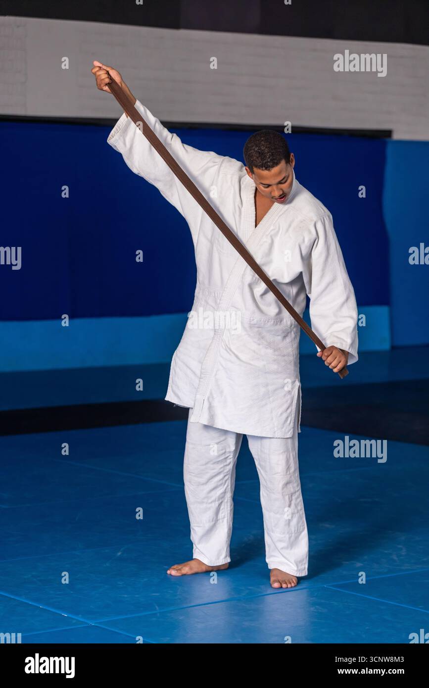 Homme afro-américain portant GI blanc tenant la ceinture brune à travers les épaules dans le gymnase avec des tapis Banque D'Images