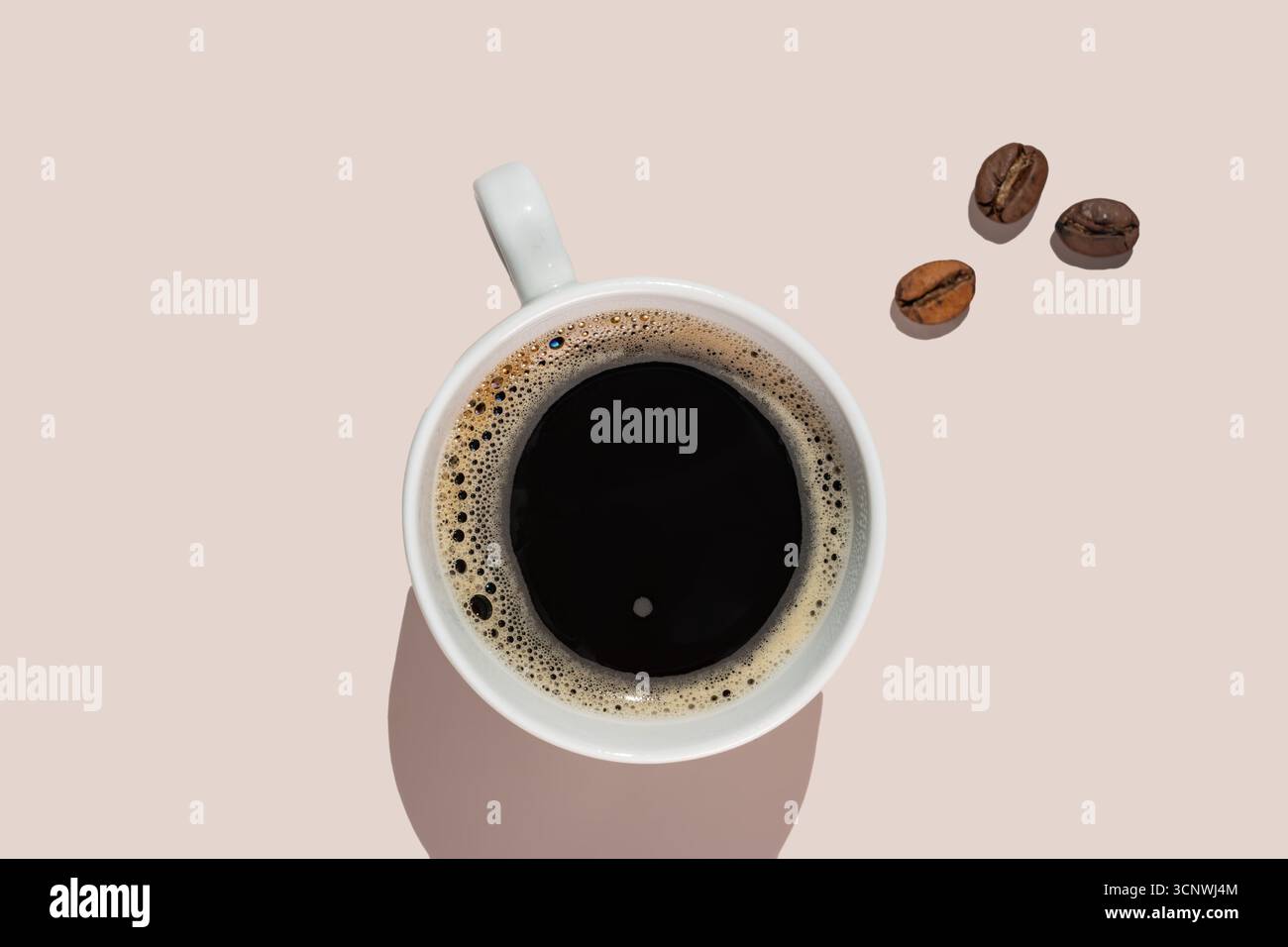 Une vue de haut en bas d'une tasse blanche remplie de café noir, accompagnée de grains de café dispersés sur un fond beige clair, créant un et minimaliste Banque D'Images