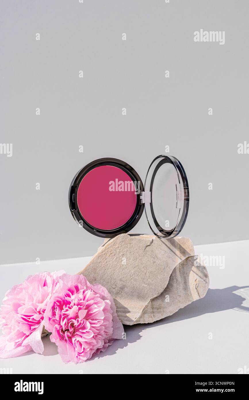 Un élégant compact de fard à joues et de poudre repose élégamment sur des pierres naturelles, accompagné de délicates fleurs roses. La scène évoque un sens du beau serein Banque D'Images