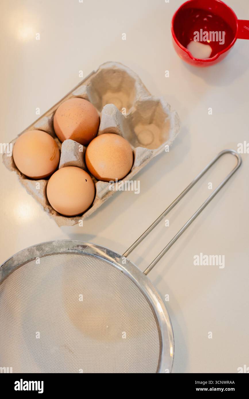 Le carton d'oeufs en carton contient quatre oeufs bruns sur le comptoir de la cuisine près de la tasse graduée et de la passoire Banque D'Images Le carton d'oeufs en carton contient quatre oeufs bruns sur le comptoir de la cuisine près de la tasse graduée et de la passoire Banque D'Images