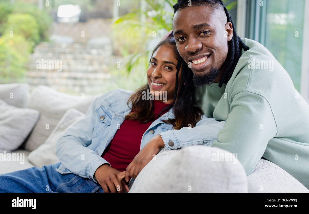 Couple diversifié assis sur un canapé gris clair dans le salon par fenêtre en verre avec plante en pot Banque D'Images