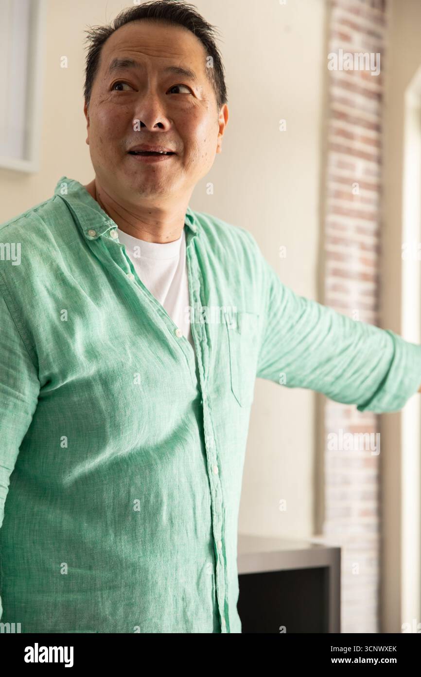 Homme asiatique faisant des gestes vers le mur d'accent exposé à l'intérieur du salon avec cadre de fenêtre blanc Banque D'Images