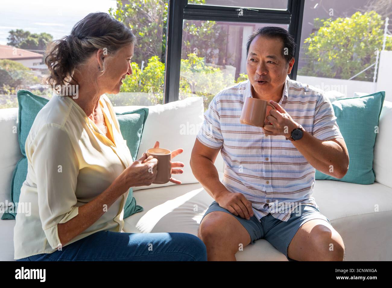 Couple diversifié sirotant du café de tasses en céramique tout en discutant sur le canapé dans le salon Banque D'Images