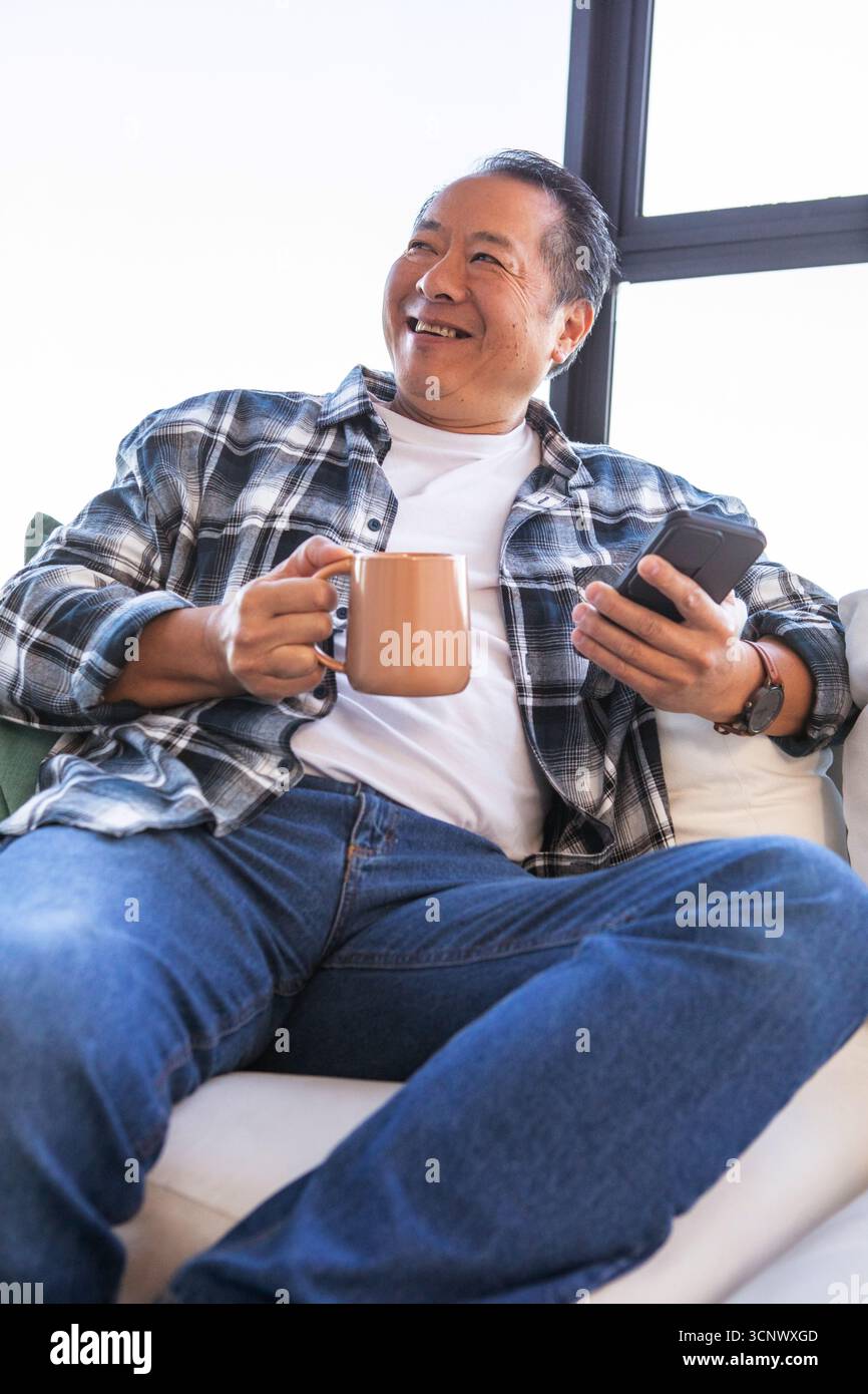 Adulte adulte chinois homme souriant tout en tenant smartphone et tasse sur le canapé par fenêtre Banque D'Images