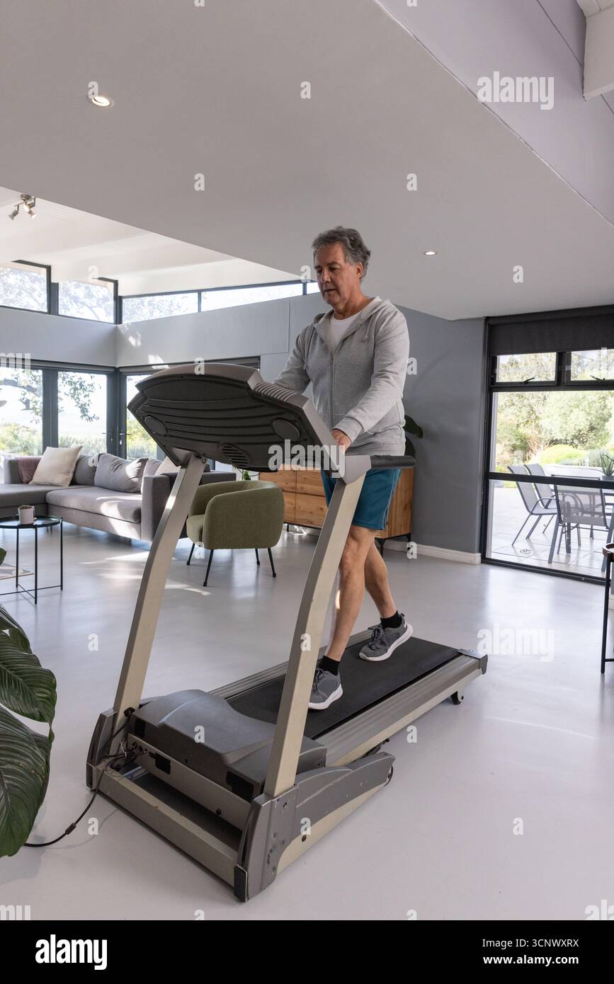 Homme senior marchant sur le tapis roulant à la maison portant des vêtements de sport avec portes coulissantes en verre, fauteuil Banque D'Images