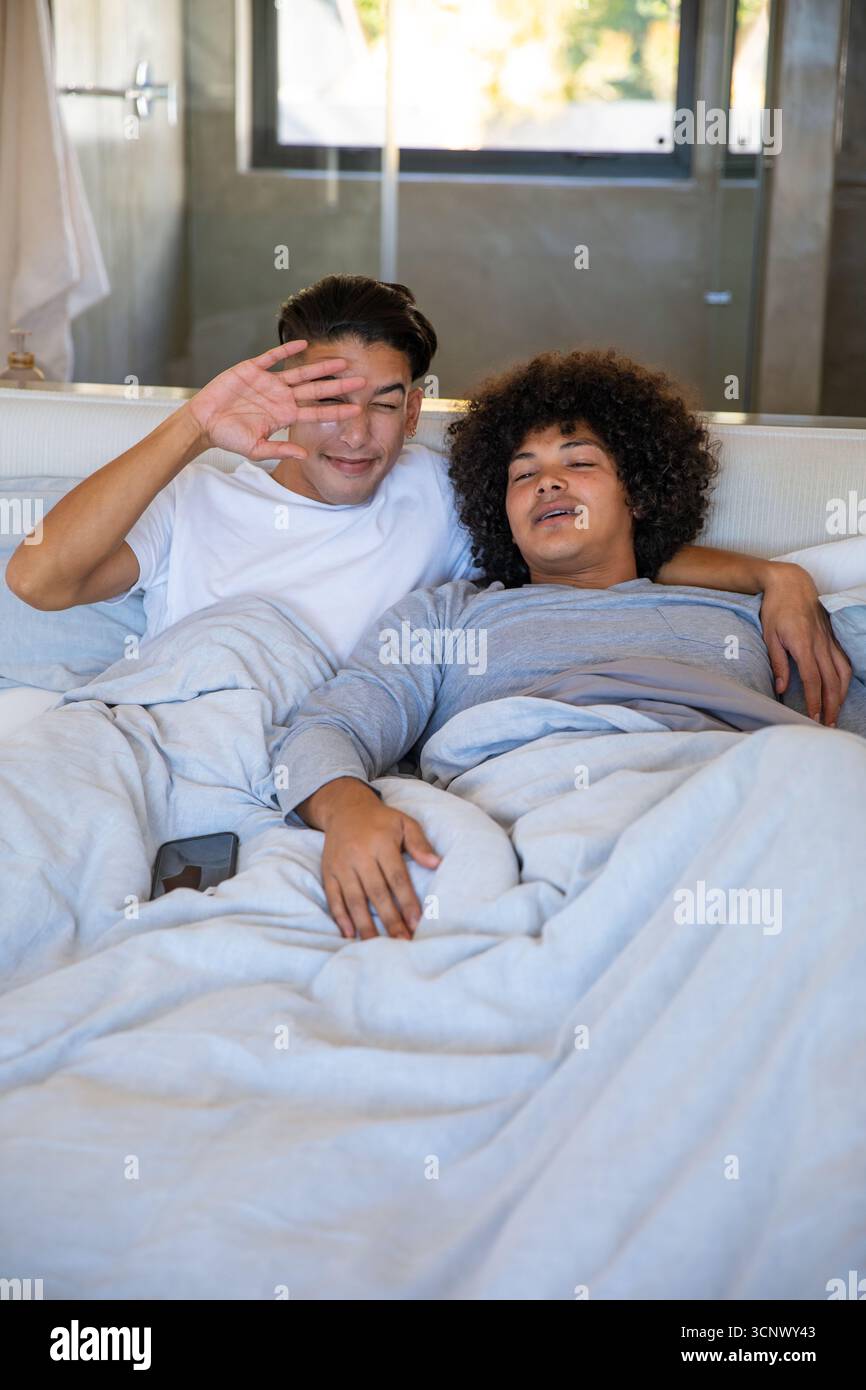 Divers partenaires masculins protégeant les yeux et câlins sous couette sur le lit dans la chambre avec smartphone Banque D'Images