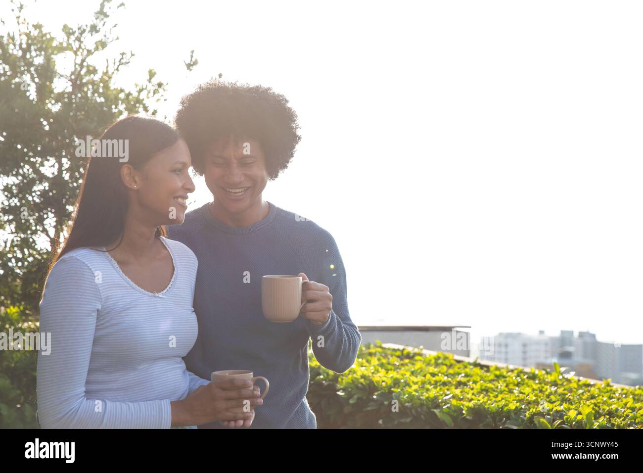 Couple diversifié tenant des tasses à café, souriant l'un à l'autre sur le toit-terrasse avec horizon de la ville Banque D'Images Couple diversifié tenant des tasses à café, souriant l'un à l'autre sur le toit-terrasse avec horizon de la ville Banque D'Images