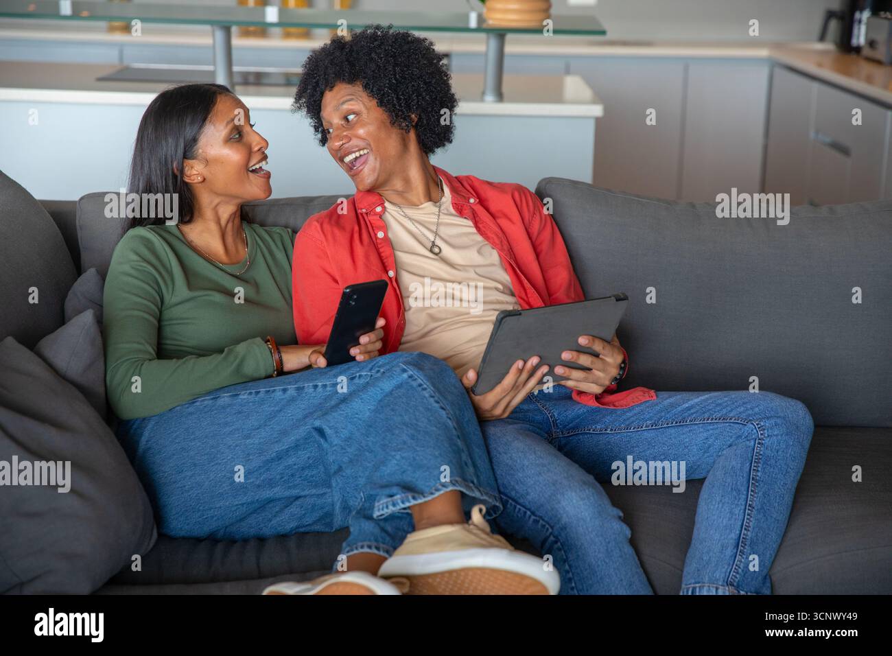 Couple afro-américain et indien parlant sur un canapé gris dans le salon à l'aide d'une tablette, smartphone Banque D'Images Couple afro-américain et indien parlant sur un canapé gris dans le salon à l'aide d'une tablette, smartphone Banque D'Images